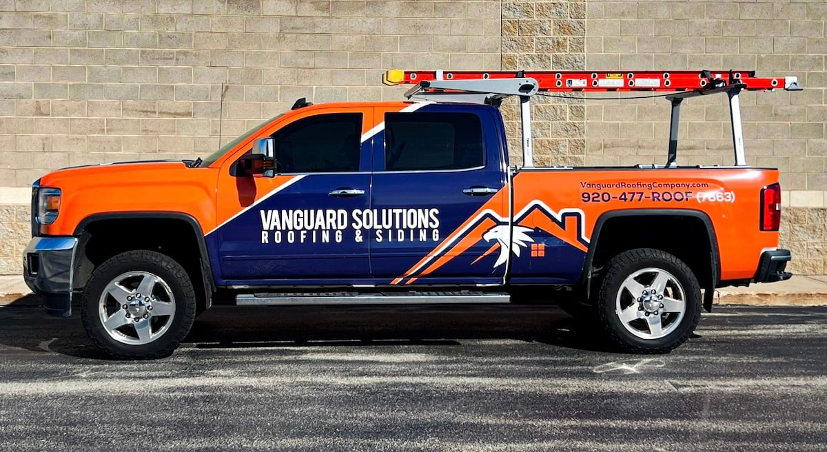 A bright orange and blue Vanguard pickup truck with a roof rack and ladder, parked by a beige brick wall.